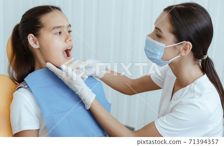 Millennial lady doctor in uniform, protective mask and rubber gloves examines of small child with open mouth Millennial lady doctor in uniform, protective mask and rubber gloves examines of small child with open mouth 71394357