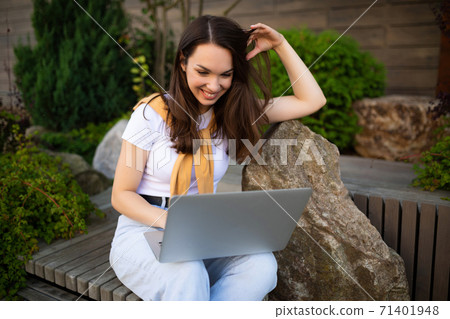cute young woman drinking coffee sits on a bench with a laptop on the street cute young woman drinking coffee sits on a bench with a laptop on the street 71401948