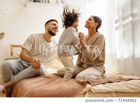 happy diverse family mom, dad and child laughing, playing and jumping in bed at home. happy diverse family mom, dad and child laughing, playing and jumping in bed at home. 71404802