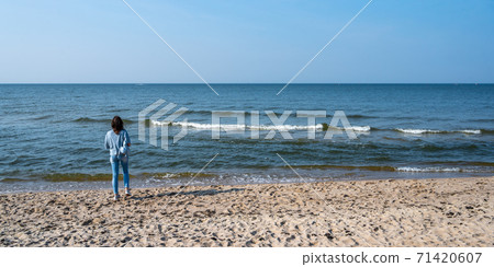 Woman in blue jeans and a blouse stands on a sandy beach and looks into the distance at sea with 71420607