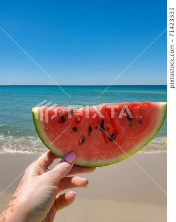 one hand holds a slice of fresh and juicy watermelon with a blurred sea and blue sky in summer 71423331