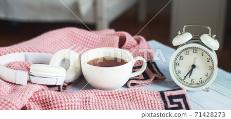 A cup of hot tea, knitted sweater, alarm clock and headphones in the bedroom. winter morning concept. banner 71428273