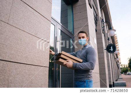 Delivery man wearing medical mask ringing door for delivery carton boxes with hot pizza. 71428650