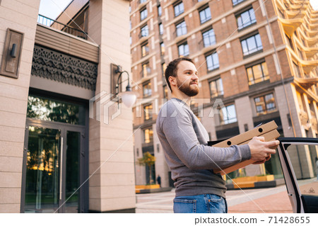Low-angle shot of delivery man standing at city street near car and holding carton boxes with food. 71428655