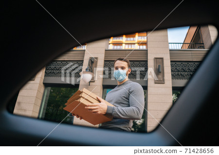 Shooting from interior car of delivery man wearing medical protective mask holding boxes with pizza. 71428656