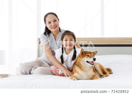 Asian mother and daughter sitting on the bed with Shiba Inu lying on the right side. Concept family with dog at home. Asian mother and daughter sitting on the bed with Shiba Inu lying on the right side. Concept family with dog at home. 71428952