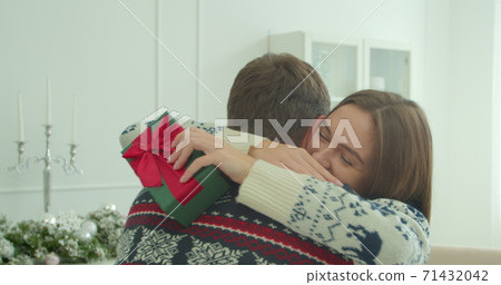 Pretty girl hugging with boyfriend at Christmas celebrating holidays together. Close up of young beautiful woman hugging man holding christmas present. Family tradition. 71432042