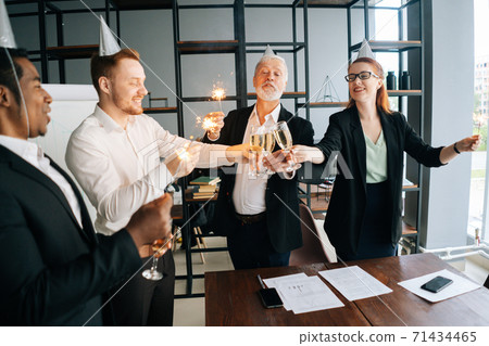Cheerful business team of multiethnic coworkers holding Bengal lights, clinking glasses of champagne 71434465