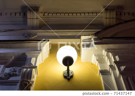 wall mount iron lantern with a white ball of plafond glow on the facade of the building with decorative architectural moulding of the cornice, close up details night scene. 71437147