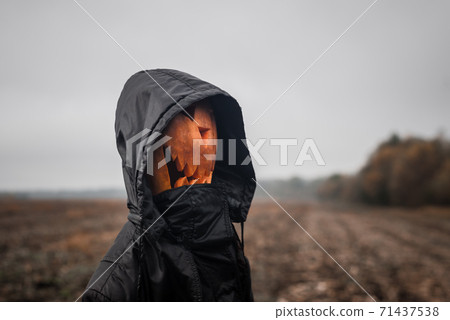 A pumpkin-headed figure Halloween jack-o'-lantern with a scary face in a black hooded robe stands in the field A pumpkin-headed figure Halloween jack-o'-lantern with a scary face in a black hooded robe stands in the field 71437538