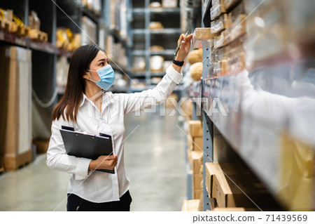 woman worker with medical mask holding clipboard and checking inventory in warehouse during coronavirus (covid-19) pandemic 71439569