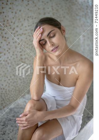 Dark-haired female wrapped in towel sitting in sauna, resting her head on her hand 71449210