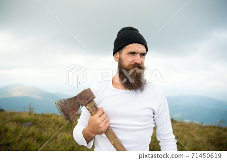 Hipster with beard on strict face holds axe, skyline on background. Lumberjack brutal Hipster with beard on strict face holds axe, skyline on background. Lumberjack brutal 71450619