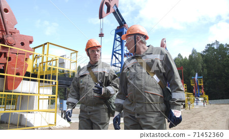 Two man oil workers walking and talking near oil pump jacks. Oil engineers overseeing site of crude oil production. Balancing drive rod pumps oil rocker. Equipment for wellhead connection oil well. Two man oil workers walking and talking near oil pump jacks. Oil engineers overseeing site of crude oil production. Balancing drive rod pumps oil rocker. Equipment for wellhead connection oil well. 71452360