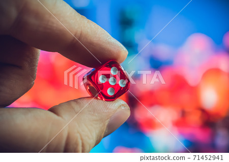 A man holds red dice in his hands between fingers against the background of night city lights A man holds red dice in his hands between fingers against the background of night city lights 71452941