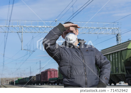 A foreman in a medical mask at a railway station looks out for trains, covering his face with his hand from the sun A foreman in a medical mask at a railway station looks out for trains, covering his face with his hand from the sun 71452942