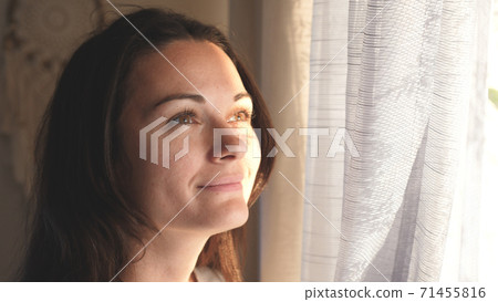 Young woman opening curtains looking through the window. Happy confident lady enjoying beautiful view and dreaming at home. Smiling girl contemplating feeling hope peaceful morning 71455816
