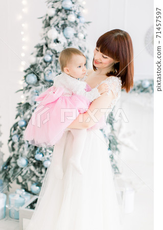 Portrait of smiling young mother holding her beautiful little baby girl, posing on the background of decorated Christmas tree. Family time, winter holidays concept Portrait of smiling young mother holding her beautiful little baby girl, posing on the background of decorated Christmas tree. Family time, winter holidays concept 71457597