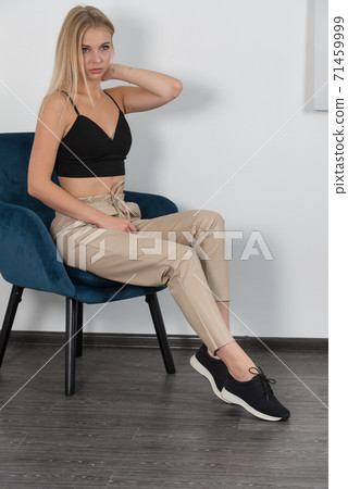 Stylish beautiful young blond woman in a tight biege pants, black top and white shirt near a white wall in the room. Attractive girl model posing. Stylish beautiful young blond woman in a tight biege pants, black top and white shirt near a white wall in the room. Attractive girl model posing. 71459999