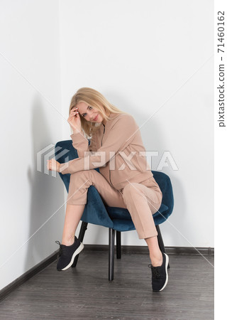 Stylish beautiful young blond woman in a biege tracksuit poses near a blue chair in the room. Stylish beautiful young blond woman in a biege tracksuit poses near a blue chair in the room. 71460162