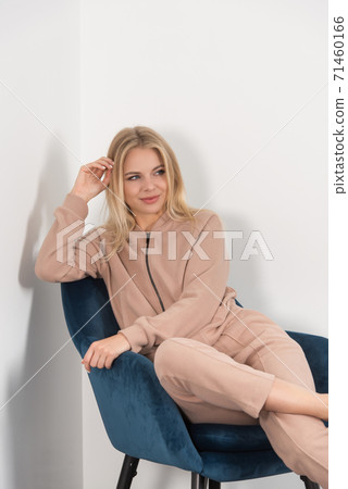 Stylish beautiful young blond woman in a biege tracksuit poses near a blue chair in the room. Stylish beautiful young blond woman in a biege tracksuit poses near a blue chair in the room. 71460166