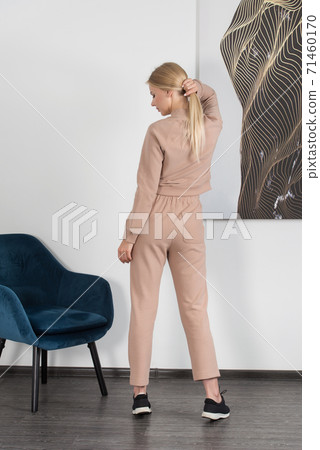 Stylish beautiful young blond woman in a biege tracksuit poses near a blue chair in the room. 71460170