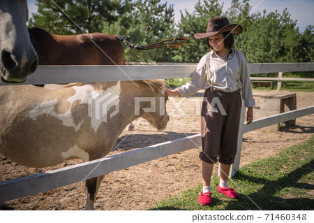 Smiling girl in cowboy clothes strokes little pony in paddock on sunny summer day. Taking care of animals concept 71460348