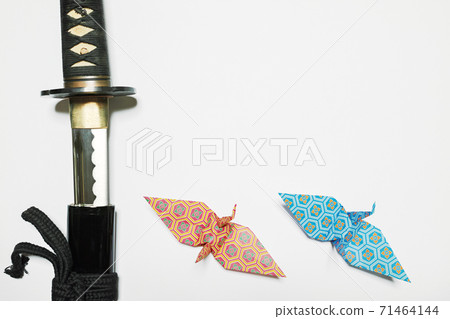 A Japanese sword placed on the left edge of white paper and a folded paper crane with an orange and blue turtle shell Hanabishi pattern placed below A Japanese sword placed on the left edge of white paper and a folded paper crane with an orange and blue turtle shell Hanabishi pattern placed below 71464144