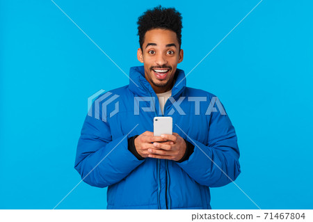 Excited happy smiling african american in padded winter jacket, holding smartphone and grinning upbeat, receive invitation party, chatting friends, got famous on social media, blue background 71467804