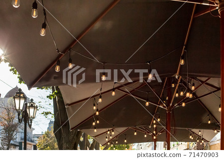 textile umbrella with a wooden frame and edison pendant lamps glowing with warm light on a backyard terrace against a background of green trees with a sky and retro street lantern, nobody. textile umbrella with a wooden frame and edison pendant lamps glowing with warm light on a backyard terrace against a background of green trees with a sky and retro street lantern, nobody. 71470903