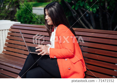 Beautiful brunette woman in red jacket runs on laptop on the internet on the street Beautiful brunette woman in red jacket runs on laptop on the internet on the street 71471552