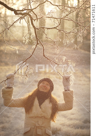 Portrait of young woman in coat, hat and mittens touching snowy icy tree branch and looking up in sunny winter forest Portrait of young woman in coat, hat and mittens touching snowy icy tree branch and looking up in sunny winter forest 71471611