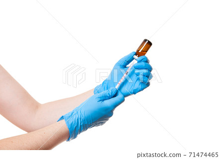 hands in blue medical gloves are holding a glass bottle with a vaccine while typing a drug into an injection syringe, medical theme close up isolated on white background with copy space. hands in blue medical gloves are holding a glass bottle with a vaccine while typing a drug into an injection syringe, medical theme close up isolated on white background with copy space. 71474465