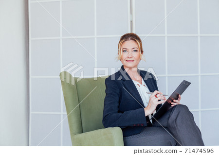 Woman in business suit at work in office with folder sits in chair Woman in business suit at work in office with folder sits in chair 71474596