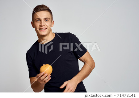A guy with an orange in his hand and in a black T-shirt on a light background cropped view A guy with an orange in his hand and in a black T-shirt on a light background cropped view 71478141