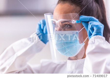 Protective shield adjustment by medical staff worker wearing white overall, blue gloves and disposable facemask Protective shield adjustment by medical staff worker wearing white overall, blue gloves and disposable facemask 71489017