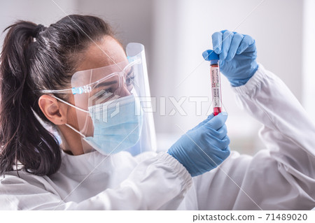 Laboratory worker wearing protective equipment examines an ampule with a Covid-19 test Laboratory worker wearing protective equipment examines an ampule with a Covid-19 test 71489020