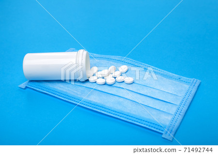 medical mask with a white plastic bottle of a medicinal product with a scattered pile of pills for the treatment of the respiratory tract of the lungs on a blue background with copy space. medical mask with a white plastic bottle of a medicinal product with a scattered pile of pills for the treatment of the respiratory tract of the lungs on a blue background with copy space. 71492744