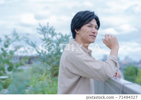 多雲的天空一個年輕的男人微笑著，一邊while著電子煙在建築物的屋頂上 71494327
