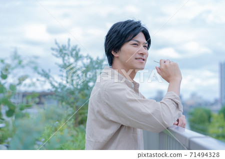 多雲的天空一個年輕的男人微笑著，一邊while著電子煙在建築物的屋頂上 71494328