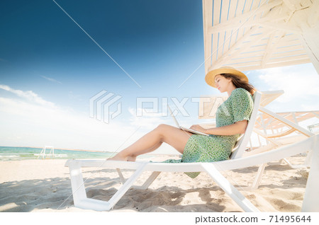 Lovely young caucasian woman freelancer in a green dress and straw hat on a schizlong under an umbrella works on her laptop. Remote work on the seaside success Lovely young caucasian woman freelancer in a green dress and straw hat on a schizlong under an umbrella works on her laptop. Remote work on the seaside success 71495644