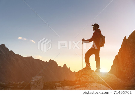 Silhouette of a man traveler in a cap with a backpack and trekking poles. Stands on the edge of a cliff against the background of mountains and the skyline in the evening at dusk Silhouette of a man traveler in a cap with a backpack and trekking poles. Stands on the edge of a cliff against the background of mountains and the skyline in the evening at dusk 71495701