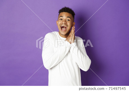 Image of excited and hopeful african-american queer guy, looking with anticipation and thrill at something, clap hands and smiling happy, standing over purple background 71497545