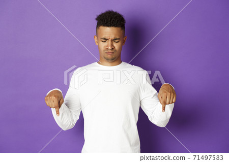 Portrait of skeptical and disappointed african-american male model in white sweatshirt, frowning and pouting upset, pointing and looking down, standing over purple background Portrait of skeptical and disappointed african-american male model in white sweatshirt, frowning and pouting upset, pointing and looking down, standing over purple background 71497583