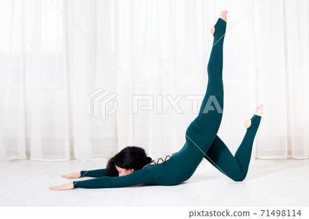 a woman in sportswear is doing a bending exercise and lifting one leg up on the floor side view of a bright gym with white curtains. 71498114