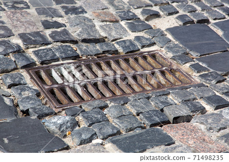 the old iron grate of the drainage system hatch on the road paved with paving stones, closeup of a sewer grate. 71498235