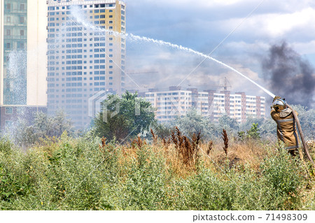 firefighter in a protective suit with a hose pours water over dry grass rescuing from a fire in the background a clearing with dry grass filled with smoke and multi-storey residential buildings. firefighter in a protective suit with a hose pours water over dry grass rescuing from a fire in the background a clearing with dry grass filled with smoke and multi-storey residential buildings. 71498309