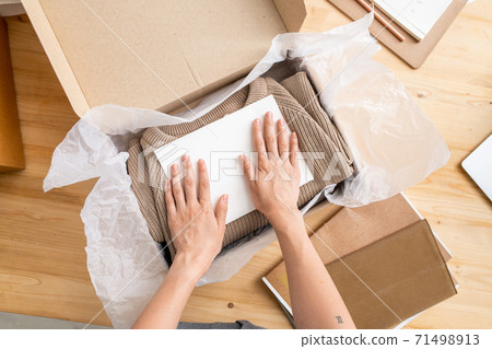 Hands of young manager of online shop putting paper on top of folded sweater Hands of young manager of online shop putting paper on top of folded sweater 71498913