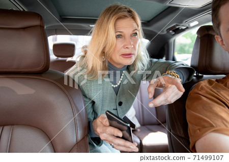 Blond elegant mature businesswoman talking to taxi driver from backseat 71499107