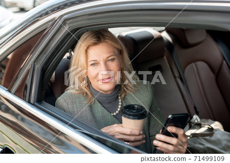 Mature woman with glass of coffee using phone while sitting on backseat of car 71499109